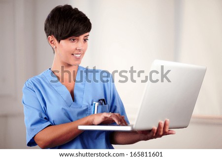 Portrait of a lovely latin doctor on blue uniform using her laptop while standing and smiling on hospital