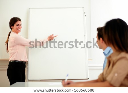 Portrait of pretty adult businesswoman pointing at whiteboard while smiling at professional colleagues on office during a conference - copyspace
