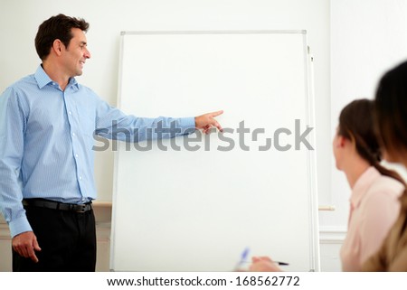 Portrait of an attractive hispanic male on blue shirt pointing and looking at whiteboard while standing in front of female team on workplace - copyspace