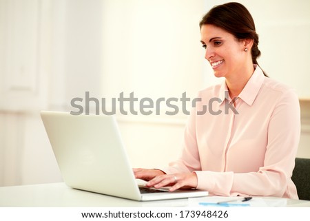 Portrait of beautiful 20s businesswoman on pink blouse using her laptop while smiling and sitting on office desk - copyspace