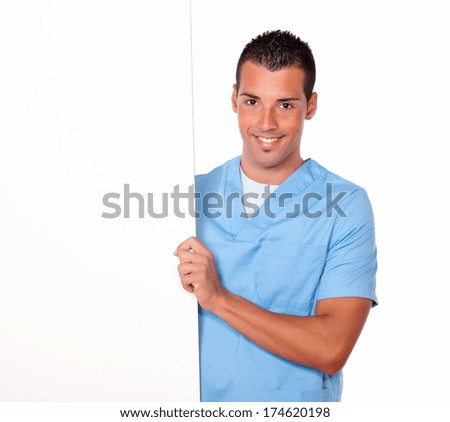 Portrait of attractive latin nurse guy on blue medical uniform holding a blank white placard while smiling at you on isolated background - copyspace