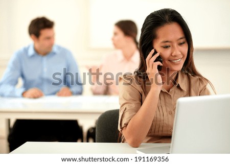 Portrait of adult asiatic woman conversing on her cellphone while smiling and sitting on office with colleagues in background