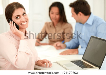 Portrait of adult businesswoman talking on her cellphone while smiling at you and sitting with male and female coworker on office desk