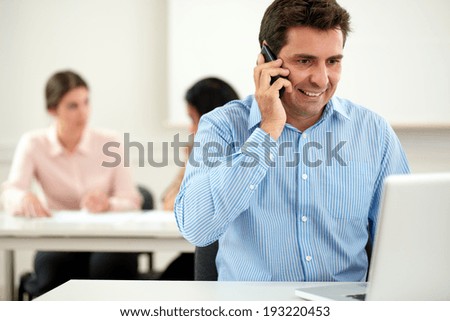 Portrait of executive latin man conversing on his cellphone while smiling and sitting on office with colleagues in background