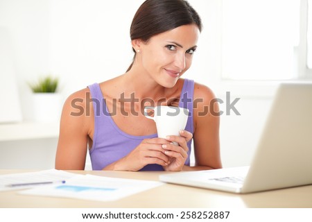 Adult woman in purple shirt sitting and surfing the web on the computer while holding a tea cup and looking at you at home