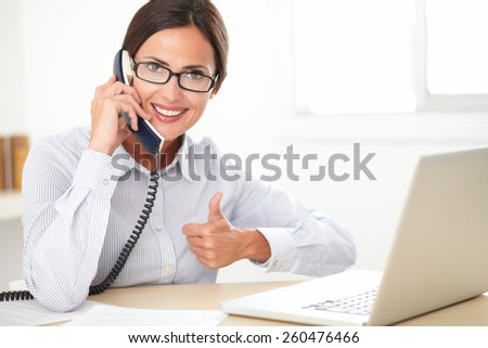 Latin receptionist with spectacles talking on the phone while using the laptop at her workplace