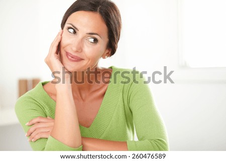 Friendly latin lady in green shirt smiling and looking satisfied to her left at home - copyspace
