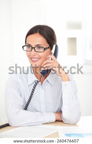Latin receptionist with spectacles conversing on the phone while smiling at her workplace