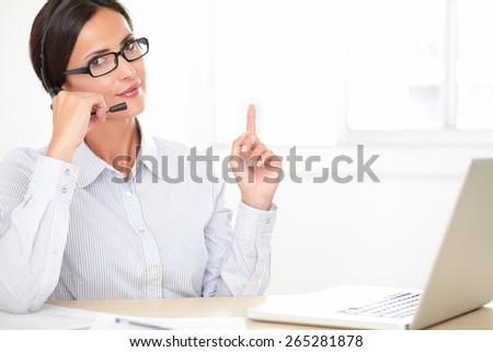 Confident secretary with glasses working on the computer on her desk while using headphones and looking at you
