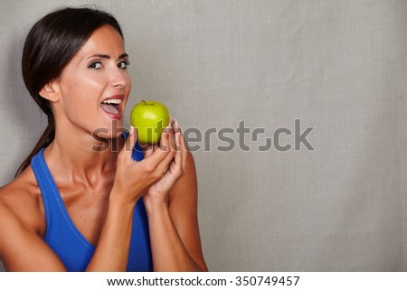 Adult female with open mouth holding apple and looking at camera on grey texture background
