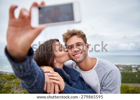 Young woman kissing her boyfriend on his cheek while taking a selfie outdoors on a nature trail