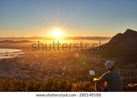 Rear view of a young guy sitting on a mountain trail watching the sunrise with the city below