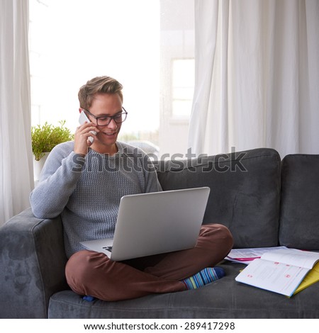 Young guy at home on his couch talking on the phone and smiling while looking at his laptop resting on his lap
