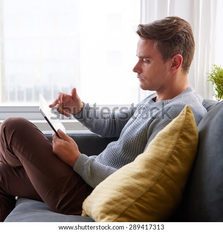 Profile shot of a guy using a digital tablet while sitting comfortably at home on his couch