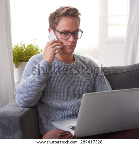 Young guy at home looking seriously at his laptop while holding a cellphone to his ear