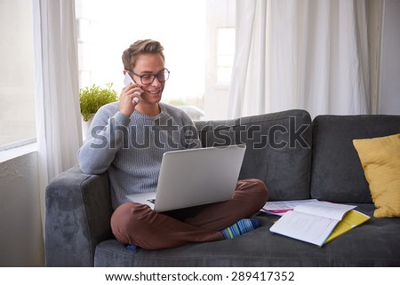 Smiling guy talking on his phone while looking at his laptop resting on his lap while he is sitting cross-legged on his couch at home