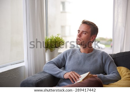 Handsome young man with a thoughtful expression pausing to think while reading a book on his couch at home
