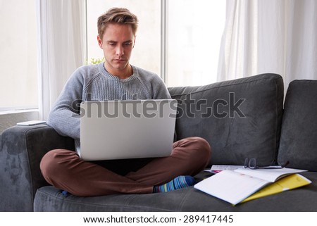 Young male student sitting cross-legged on his couch at home using his laptop while studying over the weekend