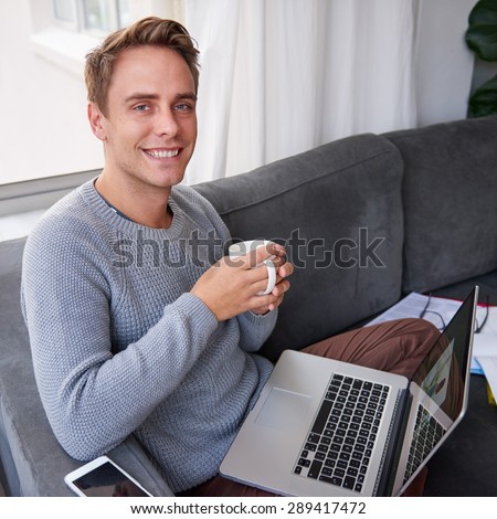 Portrait of a handsome guy sitting on his couch with his laptop open and cup of coffee in his hands looking happy and comfortable