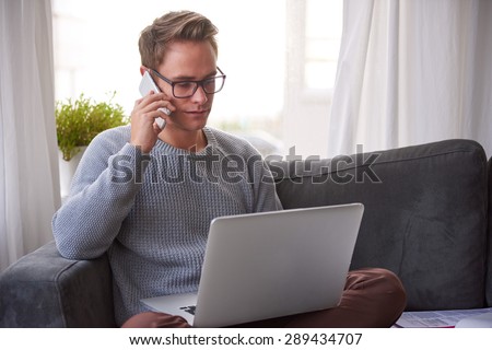 Young guy looking seriously at his laptop and listening on his phone while sitting on his couch at home
