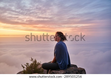 Woman sitting quietly on mountain top surrounded by serene clouds in pastel colours with early morning mist and clouds below her