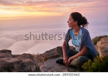 Woman sitting serenely on a mountain top at sunrise looking at the low-lying clouds and sky