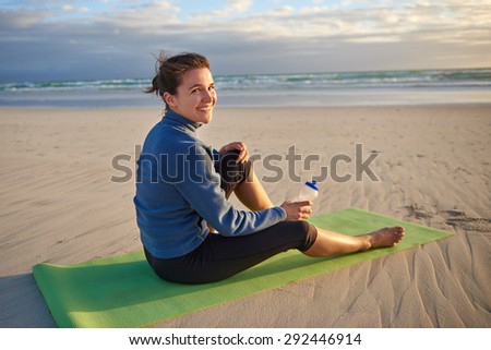 Fit and sporty young woman sitting on her yoga-mat on the beach at sunrise looking back you smiling