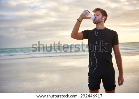 Young man looking fit and sporty drinking from his water bottle while enjoying a morning run on the beach