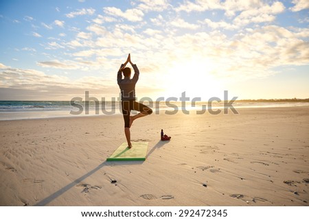 Rear view of a young woman performing the balancing yoga tree pose on a beach early in the morning at sunrise with dramatic clouds and sun flare