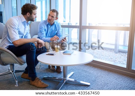 Handsome middle-aged business executive sitting with a younger coworker in a bright modern office, explaining some information to him on a digital tablet