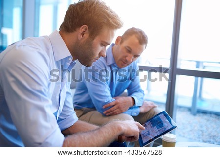 Two business executives sitting in a bright office space, looking for information together by sharing the screen of a digital tablet