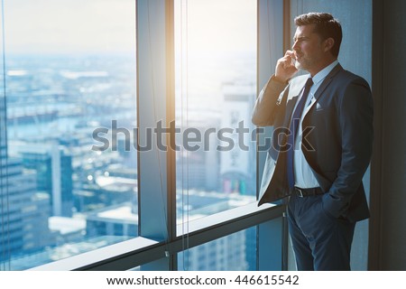 Handsome business CEO with designer stubble, talking confidently on his mobile phone while looking out of large windows in a top floor office at the city below