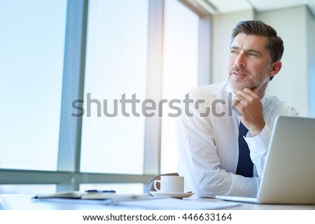 Attractive mature business executive with a stylish short beard, sitting at his office desk and looking out of his window with a thoughtful and optimistic expression