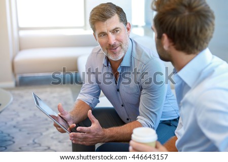 Handsome and mature corporate leader sitting in a business lounge with a young man, offering mentoring advice to him while holding a digital tablet