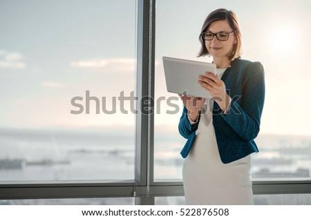 Attractive businesswoman using a digital tablet while standing in front of windows in an office building overlooking the city