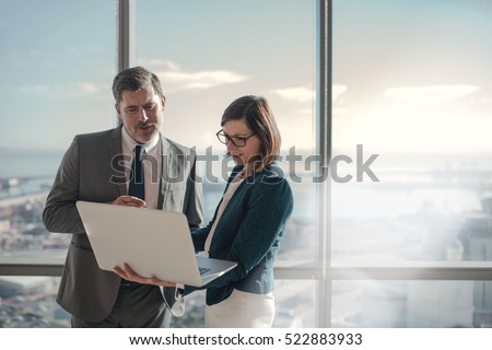 Businessman and businesswoman using a laptop together while standing in front of office building windows overlooking the city