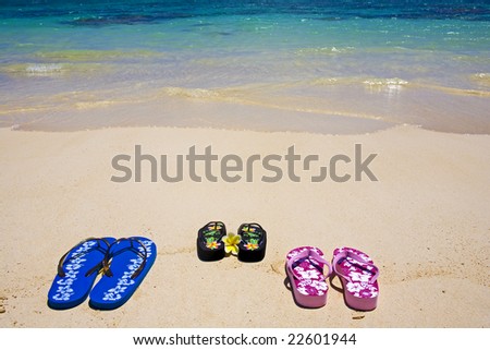 three pairs of sandals sit on the sand at water\'s edge by a lagoon in Hawaii