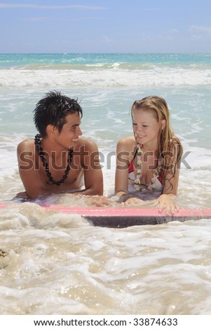 A young Hawaiian man and a blond girl visiting the islands lie on a boogie board by the ocean at the beach in Kailua talking to each other