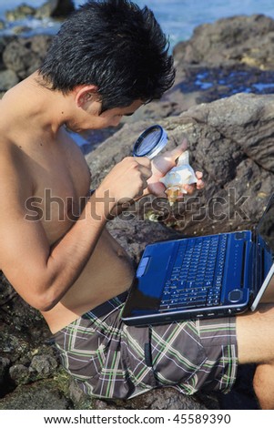 young asian man studies a seashell with his magnifying glass and computer at a tidepool
