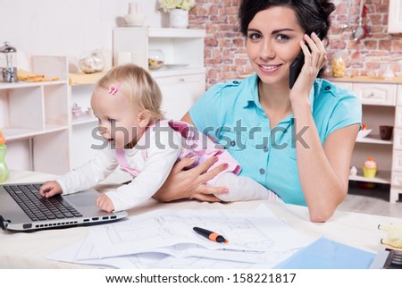 Young business woman with baby in the kitchen working with laptop, speaks by phone