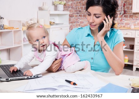 Young business woman with baby in the kitchen working with laptop, speaks by phone