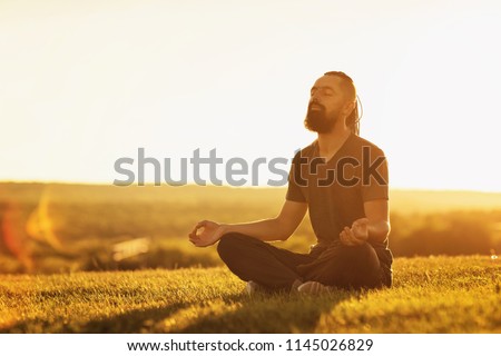 Bearded man is meditating in lotus yoga pose at summer outdoor sunset background sitting at a golden field.