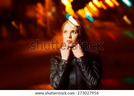 A beautiful glamour girl in jacket and muffler is standing in the city street at colored flares background.