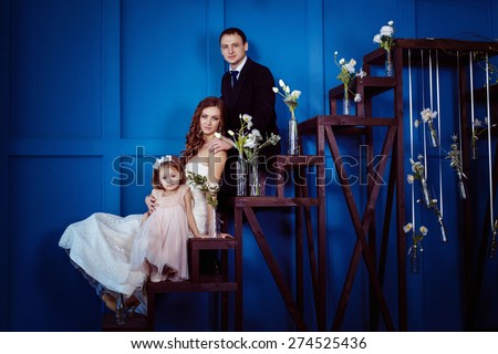 A happy family of beautiful pregnant mother in wedding dress, cheerful father and cute little girl are sitting on a ladder with spring flowers at a blue wall background.