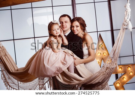 A happy family concept. Beautiful pregnant wife in wedding dress,  her cute daughter and young father are standing near a hammock at a window background.