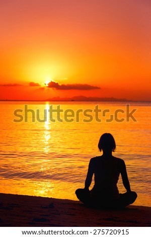 A silhouette of meditating woman at the sea shore at orange sunset background.