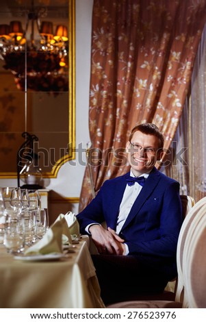 Elegant dressed cheerful businessman in glasses is sitting in luxury restaurant waiting for dinner in good mood.