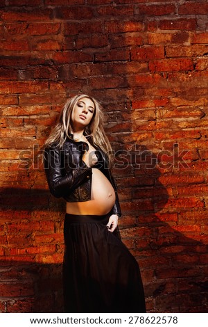 A beautiful pregnant woman in skirt and jacket is standing at a textured bricks wall with shadows on it.