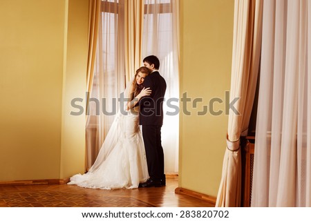 Beautiful elegant wedding couple is embracing tenderly in arch at curtained window and yellow walls background in a stunning vintage hall. Love and happy family concept.