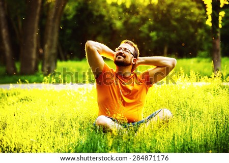 A happy smiling pleased man is sitting on green grass and folding arms behind head at sunny summer day at park background. Concept of wellbeing and healthy lifestyle
